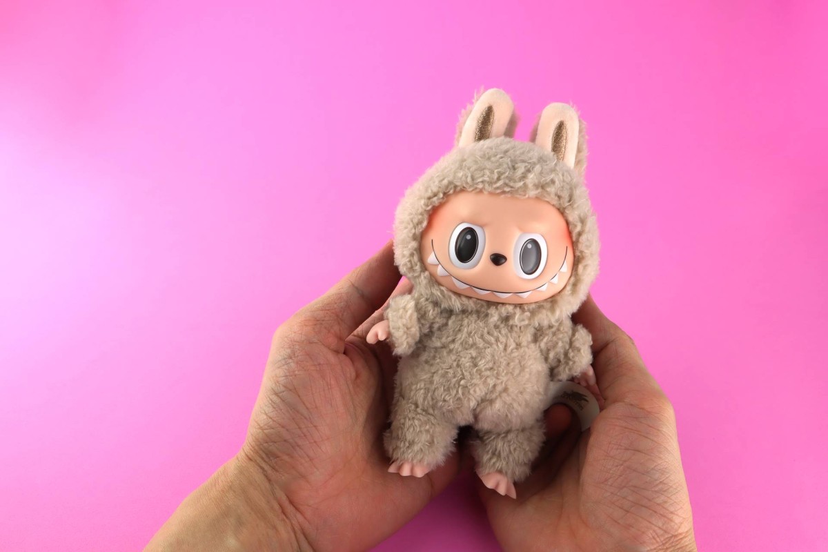 A plush toy in a fuzzy bunny suit held in two hands against a pink background