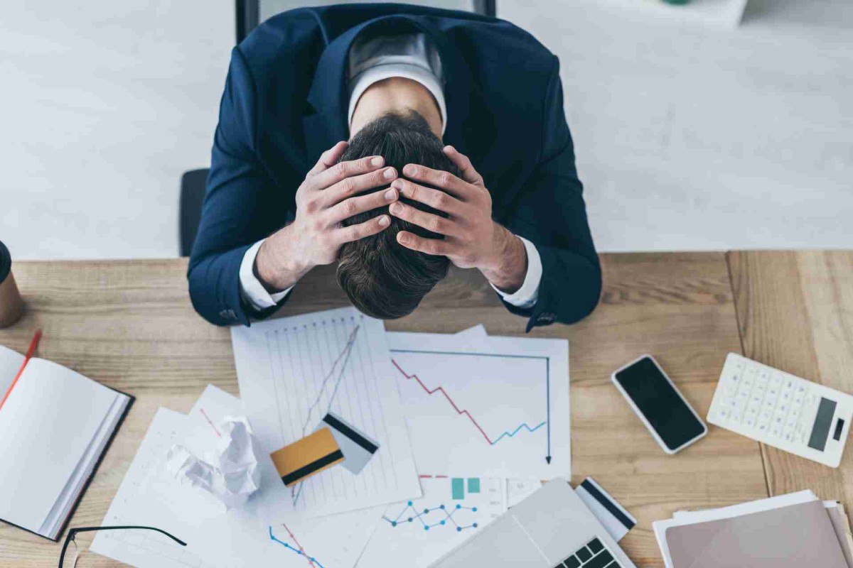 Frustrated businessman sitting at a desk with head in hands, surrounded by financial charts and a laptop