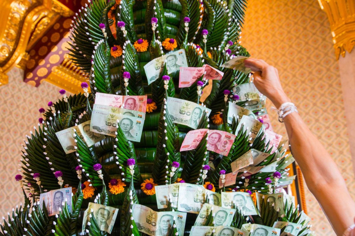 Person placing Thai banknotes on a decorated money tree with green leaves and colorful flowers