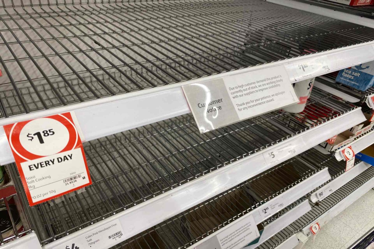 Empty supermarket shelves where cooking salt is sold, with only a few products remaining and a sign explaining the item is out of stock