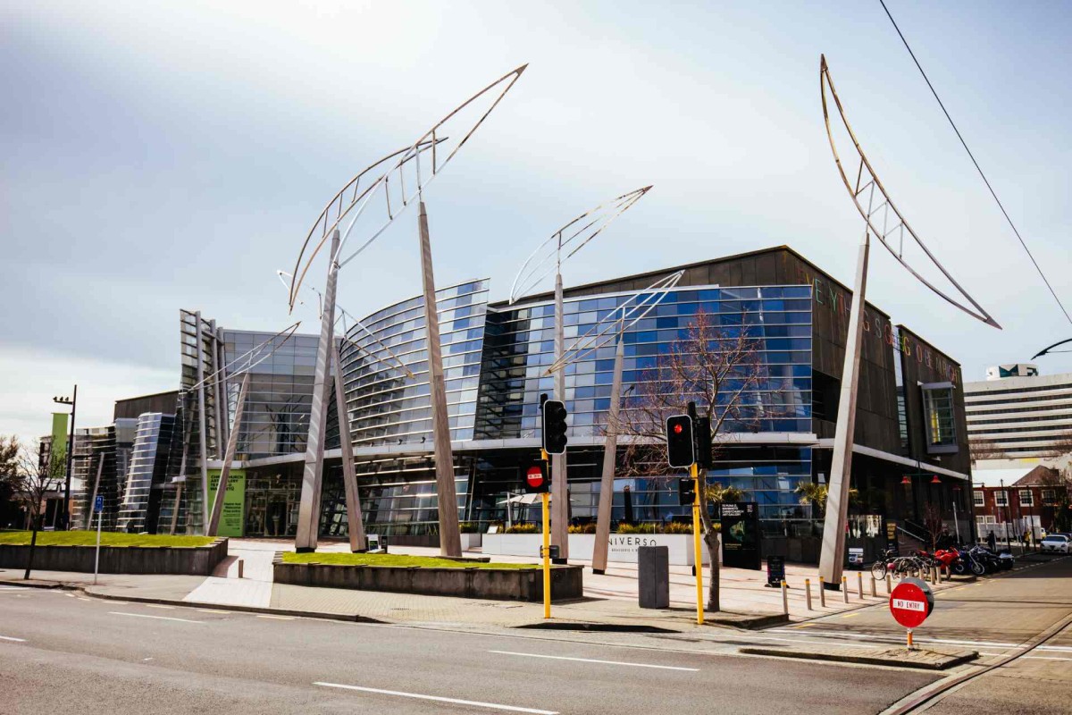 Contemporary glass-fronted building with curved facade and tall metal sculptures on a street corner with traffic lights