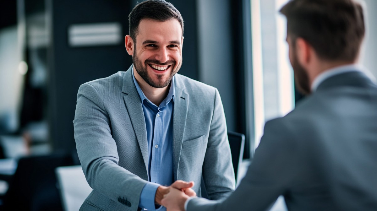 A smiling man in a suit shakes hands with another man during a professional meeting