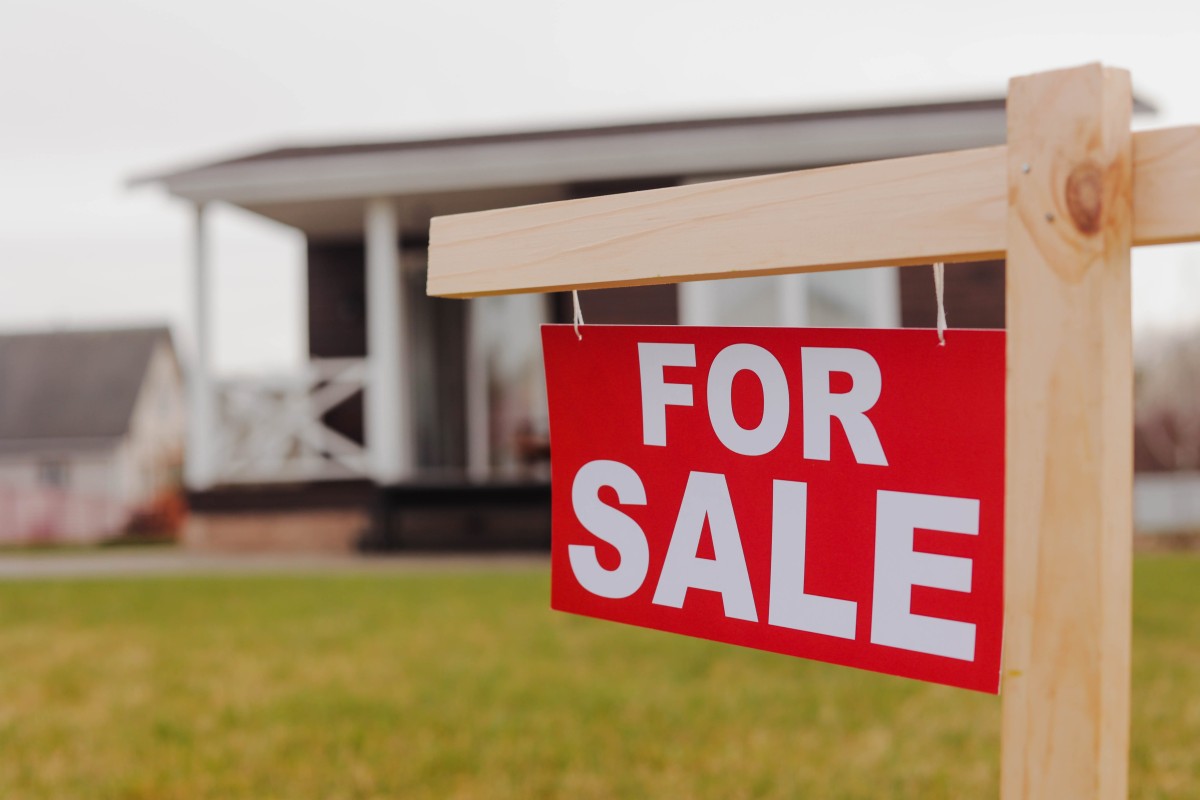 Red “For Sale” sign hanging on a wooden post in front of a house