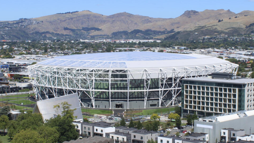 A modern stadium with a white structural frame and transparent roof, surrounded by buildings and hills in the background.