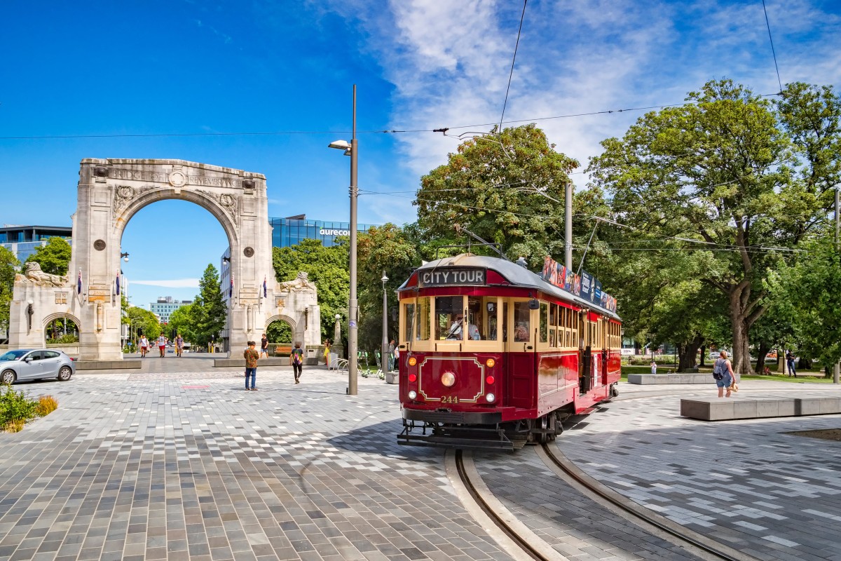 Red vintage tram passing the Bridge of Remembrance in a sunny city square with trees and pedestrians