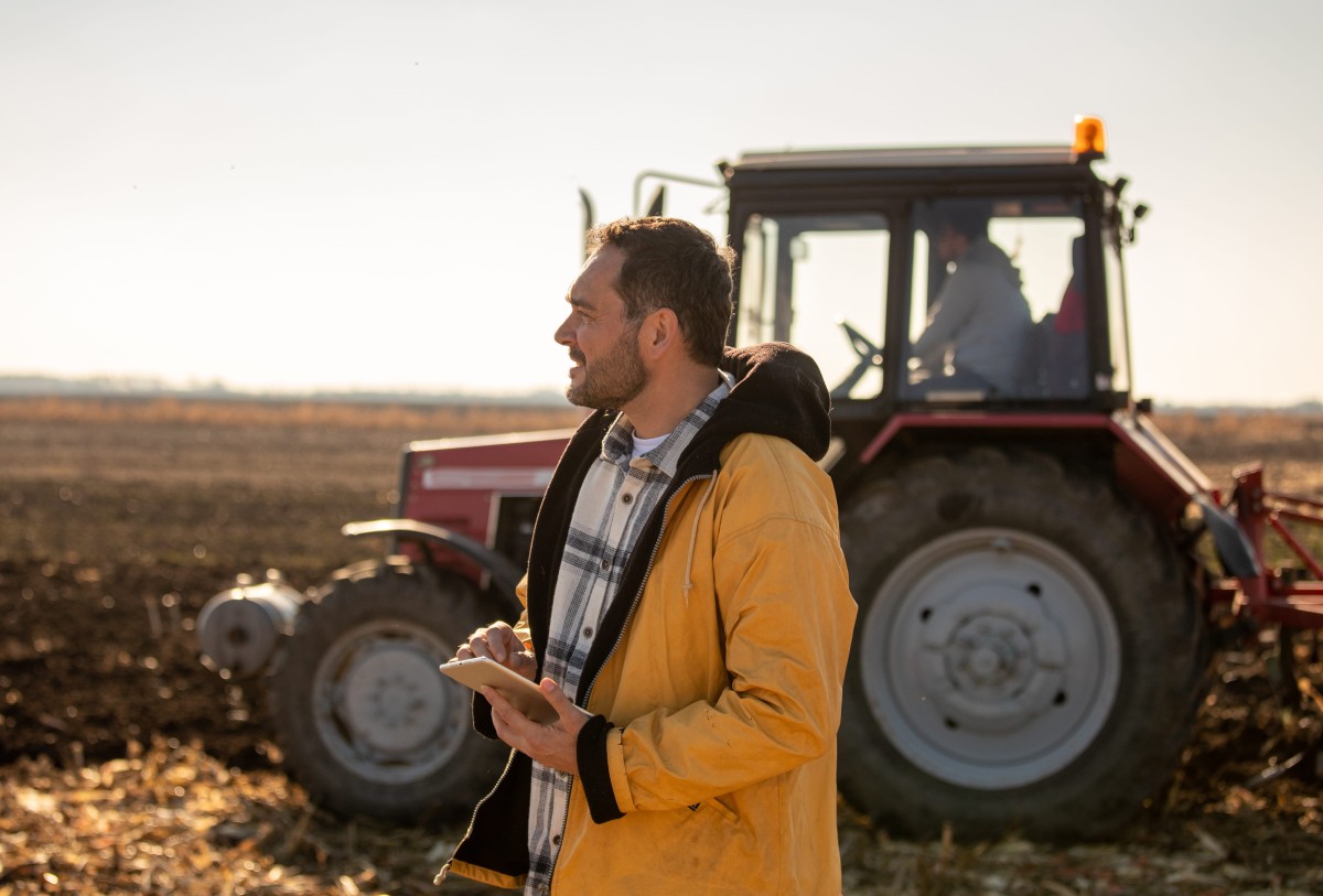 A man in a yellow jacket holding a tablet stands in a field beside a tractor.