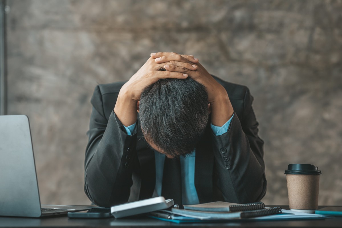 Stressed man holding head at desk during workplace crisis with laptop and paperwork