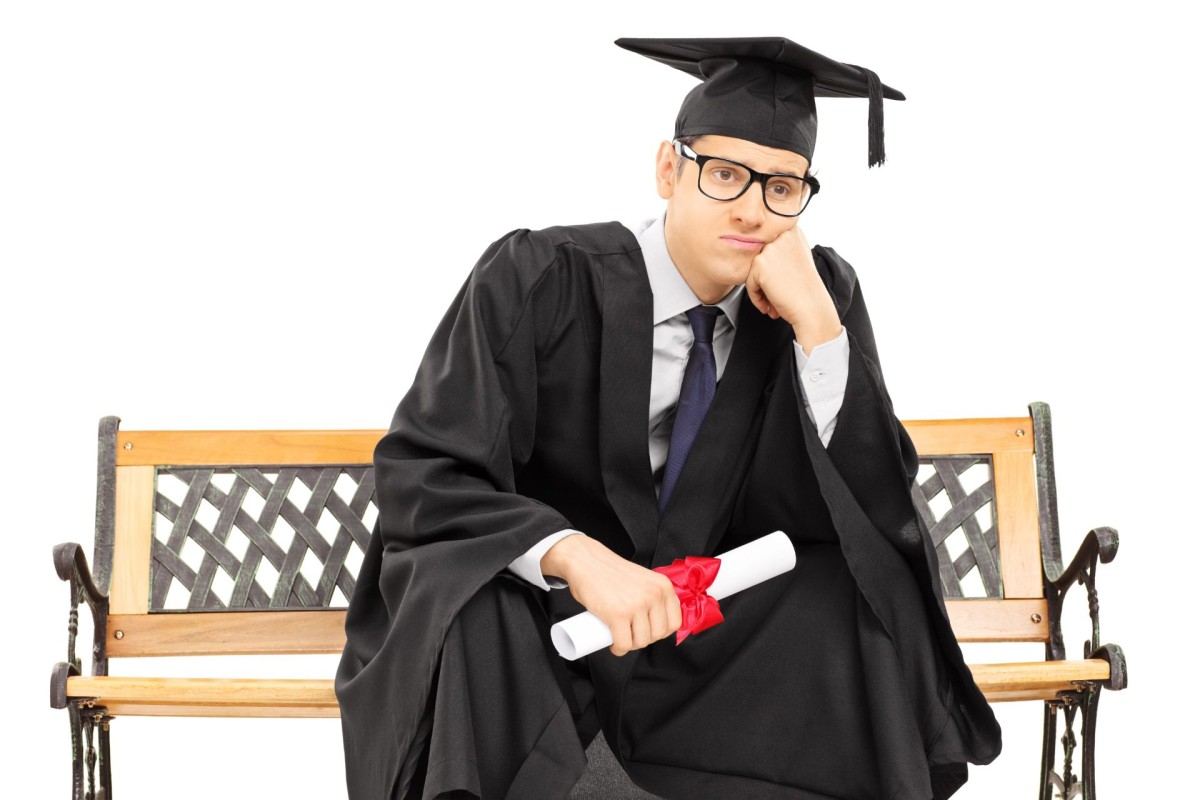 A young man in a graduation cap and gown sitting on a bench with a bored or discouraged expression, holding his diploma.
