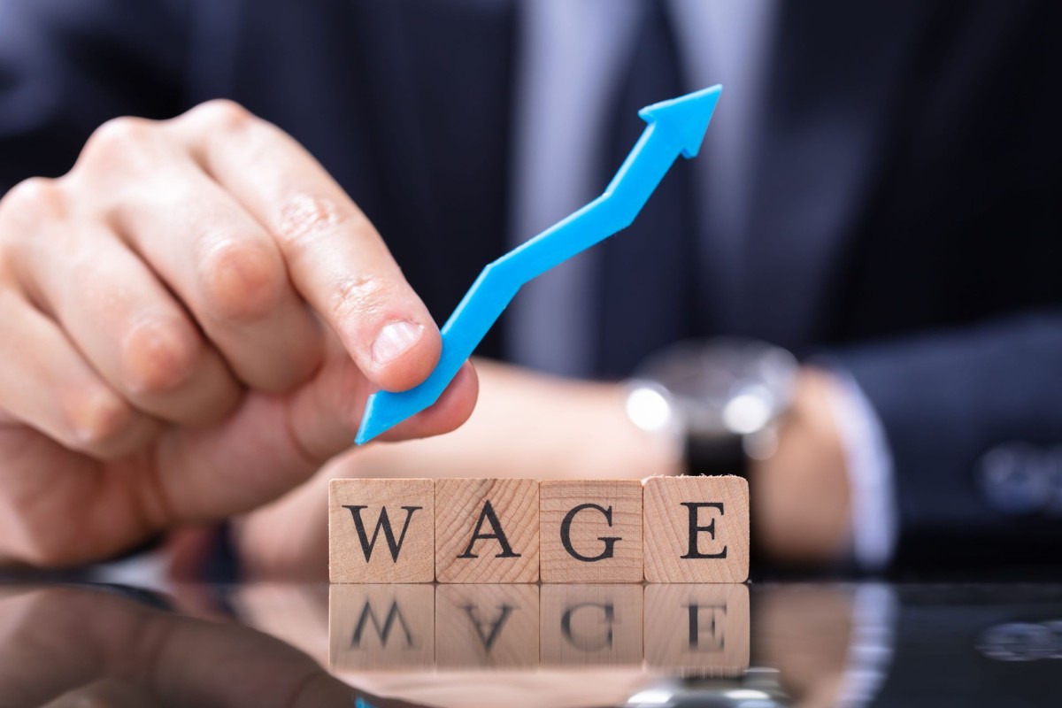Hand placing an upward arrow above wooden blocks spelling ‘WAGE’, symbolising wage growth or salary increase.