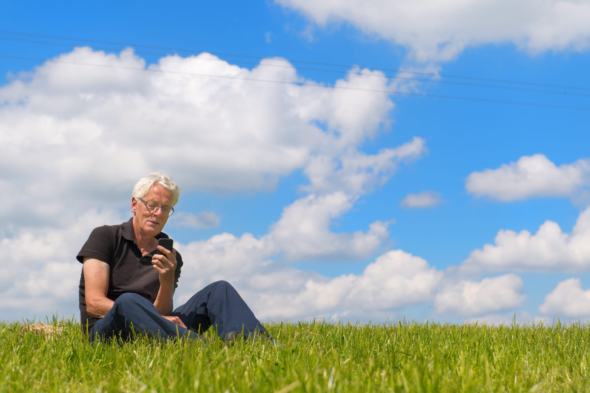 An older man sitting in grass using a smartphone under a bright blue sky