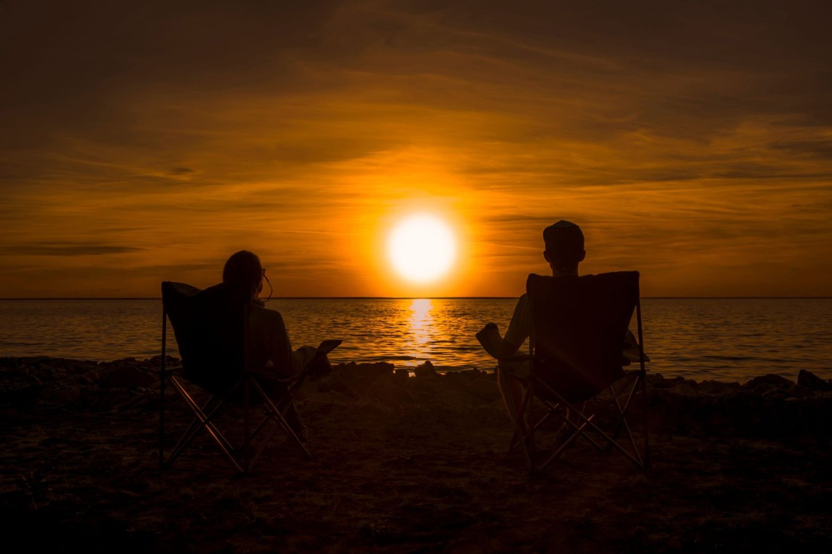 Two people sitting in chairs together by the water, watching the sunset, representing support, connection and wellbeing.