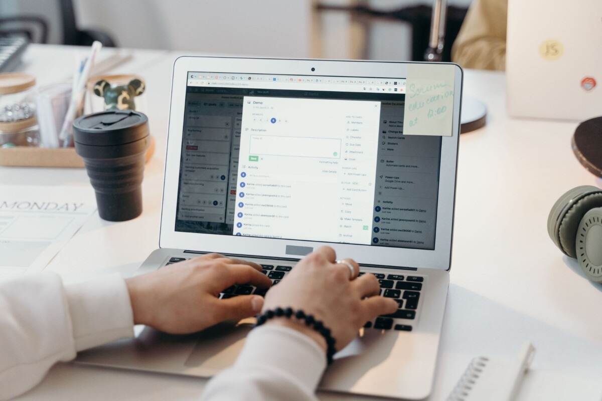 Person typing on a laptop displaying a Trello project management board, with a takeaway coffee cup, headphones and a sticky note on the desk.